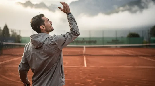 Joueur de tennis en action portant une veste déperlante légère sur un court de terre battue sous une bruine fine